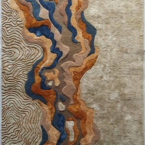 Puede incluir: Una alfombra rectangular con superficie texturizada que combina tonos azul profundo, naranja quemado, beige y arena. El dise&ntilde;o incorpora l&iacute;neas onduladas y org&aacute;nicas en el lado izquierdo y una secci&oacute;n beige s&oacute;lida y suave a la derecha.
