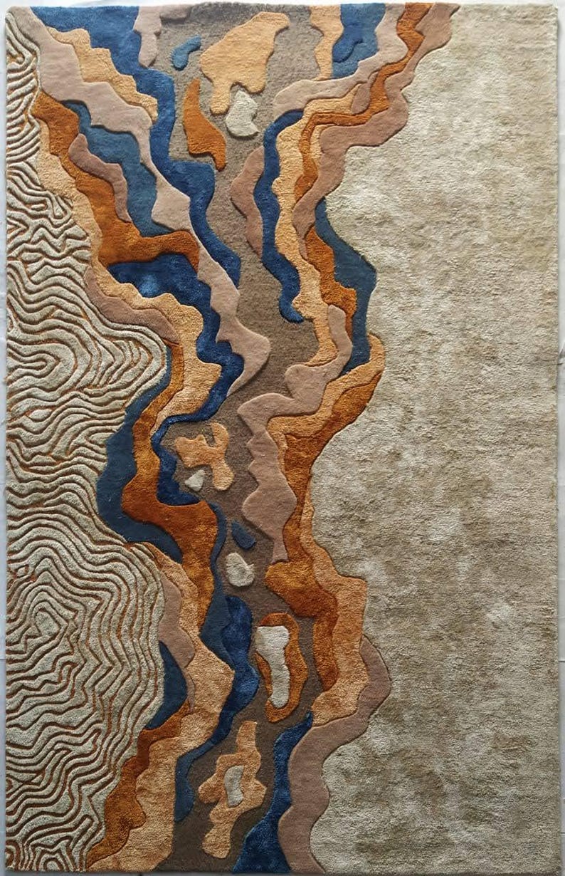 Puede incluir: Una alfombra rectangular con superficie texturizada que combina tonos azul profundo, naranja quemado, beige y arena. El dise&ntilde;o incorpora l&iacute;neas onduladas y org&aacute;nicas en el lado izquierdo y una secci&oacute;n beige s&oacute;lida y suave a la derecha.