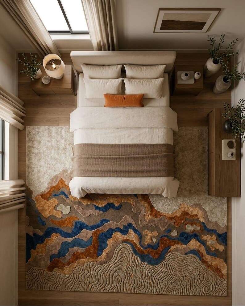 Puede incluir: Un dormitorio con una cama de ropa de cama color crema, una manta marr&oacute;n y un coj&iacute;n lumbar naranja. El suelo est&aacute; cubierto por una alfombra grande con un patr&oacute;n de paisaje abstracto y texturizado en tonos azul, marr&oacute;n, beige y crema.