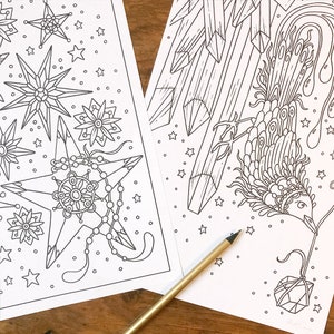 May include: Two detailed black and white coloring pages featuring celestial and geometric designs. One page showcases starbursts and star shapes, while the other depicts a bird-like figure and crystal formations. A gold pencil rests on the pages.