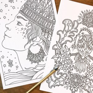 May include: Two black and white coloring pages with intricate designs. One features a profile of a woman with ornate headwear and earrings, the other a detailed floral and bird illustration. A gold pencil rests on the pages.