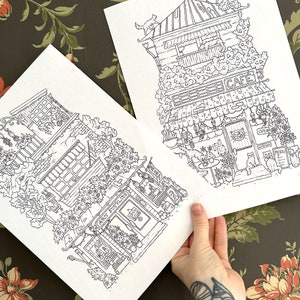 Puede incluir: Dos dibujos en blanco y negro de edificios de fachada. El primer edificio es una cafeter&iacute;a con la palabra "CAFE" sobre la puerta. El segundo edificio es una tienda con las palabras "GROCER & SUPPLIES" sobre la puerta.