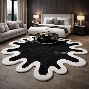 Op de afbeelding: Een zwart-wit abstract vloerkleed met een unieke, organische vorm. Het kleed heeft een zwart midden met een witte, golvende rand. Het kleed ligt in een slaapkamer met een bed, een bank en een salontafel.