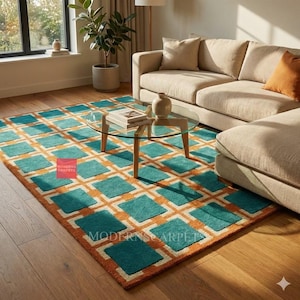 May include: A rectangular area rug with a geometric pattern in teal and orange. The rug features a grid of teal squares outlined in orange, with a cream-colored border. The rug is placed on a hardwood floor, with a beige sofa and glass coffee table in the background.