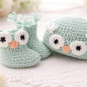 May include: A matching set of baby booties and a hat, both crocheted in a soft, light blue yarn. The booties and hat are adorned with owl faces, featuring white eyes with black centers and peach-colored beaks. The booties have a height of approximately 3 inches.