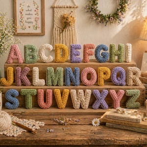 May include: A full alphabet of colorful, crocheted letters displayed on a wooden surface. Each letter is a different color, including pink, green, yellow, and blue. The letters are arranged in three rows, with a few decorative items in the background.