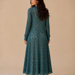 May include: A teal-colored, long-sleeved, open-front crochet cardigan. The cardigan features a delicate, openwork design and a floor-length skirt. The garment is worn over a white dress, creating a layered effect. The sleeves have a ribbed cuff.
