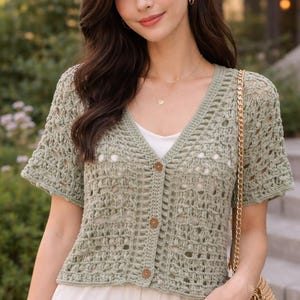 Może przedstawiać: Jasnozielony, krótki kardigan z szydełka z dekoltem w serek i drewnianymi guzikami. Kardigan jest noszony na białym topie. Złota torba na ramię wisi na ramieniu.