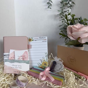 Könnte beinhalten: Ein Geschenkset mit einem rosa Notizbuch mit Hardcover, Notizblöcken mit inspirierenden Zitaten, dekorativem Washi-Tape und einem Set bunter Textmarker, die mit einem rosa Band zusammengebunden sind. Ein kleiner Geschenktüte und eine Karte sind ebenfalls enthalten.