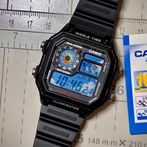 Puede incluir: Reloj digital Casio negro con esfera rectangular que muestra la hora 10:46:21. El reloj tiene hora mundial, alarmas e iluminador. La correa negra es visible, y una etiqueta indica una batería de 10 años. Mide 148 mm x 210 mm.