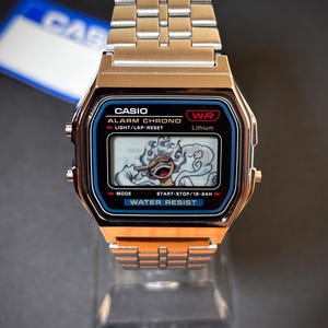 Könnte beinhalten: Eine silberfarbene Digitalarmbanduhr mit Metallgliederarmband. Das rechteckige Zifferblatt trägt die Aufschrift CASIO, ALARM CHRONO, WR und WATER RESIST. Das digitale Display zeigt eine grafische Illustration einer lachenden Figur mit lockigem Haar.