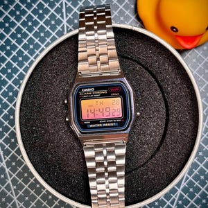 Op de afbeelding: Een zilveren Casio digitale horloge met een rechthoekige wijzerplaat die de tijd 14:49:29 weergeeft. Het horloge heeft een roestvrijstalen band en is waterbestendig. Het horloge ligt op een zwart oppervlak met een gele rubberen eend op de achtergrond.