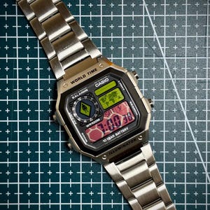 Pode incluir: Um relógio digital Casio prateado com um mostrador retangular preto e uma pulseira de metal a condizer. O mostrador do relógio mostra a hora, a data e várias funções. O relógio tem o texto "WORLD TIME" e "CASIO" no mostrador.