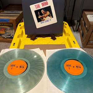 May include: An album by The Who, titled "Who Done It," with a photo of a band member. Two translucent blue vinyl records are visible, each with an orange label that reads "Side 1". The records are approximately 30 cm in diameter.