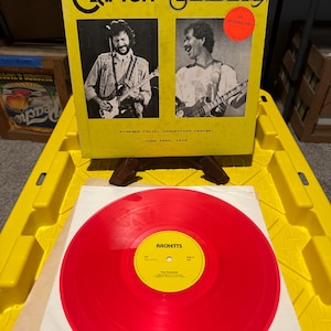 May include: A vintage vinyl record album featuring Eric Clapton and Santana. The album cover is yellow with black text and images of the musicians. A red vinyl record is visible below the album cover.