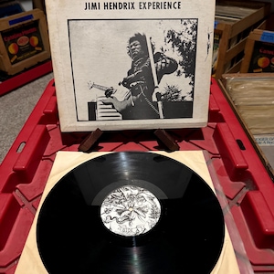 Könnte beinhalten: Eine schwarze Schallplatte mit einem schwarz-weißen Label in einer beigefarbenen Papierhülle. Das Albumcover zeigt ein Schwarz-Weiß-Bild von Jimi Hendrix und den Text "INCIDENT AT RAINBOW BRIDGE MAUI, HAWAII JIMI HENDRIX EXPERIENCE".