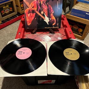 May include: A collection of vinyl records featuring a Jimi Hendrix album cover with the text "Jimi Hendrix" and "The Lord of the Strings." Two black vinyl records are in the foreground, labeled "Side One" and "Side Two."