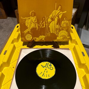 May include: A vinyl record and album cover with the text "WHO is THIS?" in yellow. The album cover features a band in sepia tones. The record label is yellow with the same text. The record is black.