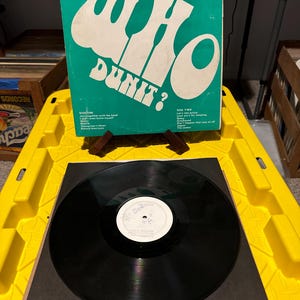 May include: A vintage vinyl record album titled "Who Dun It?" with a teal cover and white lettering. The record is black and sits on a black sleeve. The album cover is propped up on a wooden stand.