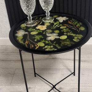 Puede incluir: Una mesa auxiliar de metal negro con una tapa redonda con un diseño botánico con flores blancas y amarillas, hojas verdes y resina transparente. Dos copas de vino claras y estampadas están sobre la mesa. La mesa tiene un marco de metal negro.