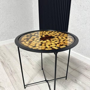 Puede incluir: Una mesa auxiliar redonda con estructura y patas de metal negro. La tapa es transparente, mostrando una colección de corchos de vino. La mesa tiene aproximadamente 50 cm de diámetro y 50 cm de altura.