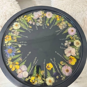Peut inclure: Un plateau rond noir orné de fleurs séchées. Les fleurs, aux tons de jaune, de violet et de blanc, sont disposées sur le bord du plateau. Le plateau est probablement en résine ou dans un matériau similaire, à des fins décoratives.