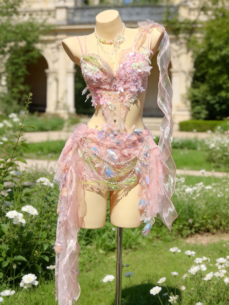 May include: A pink fairy costume displayed on a mannequin. The costume features a bodice and skirt adorned with floral and feather accents. A sheer, flowing fabric drapes from the shoulder. The costume is embellished with beads and sequins, creating a whimsical and ethereal look.