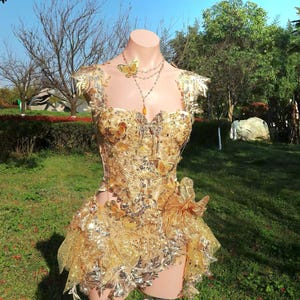 May include: A golden, embellished bodice and skirt ensemble, adorned with butterfly accents and shimmering details. The outfit is displayed on a mannequin, showcasing its intricate design and textures. A delicate necklace with a butterfly pendant completes the look.