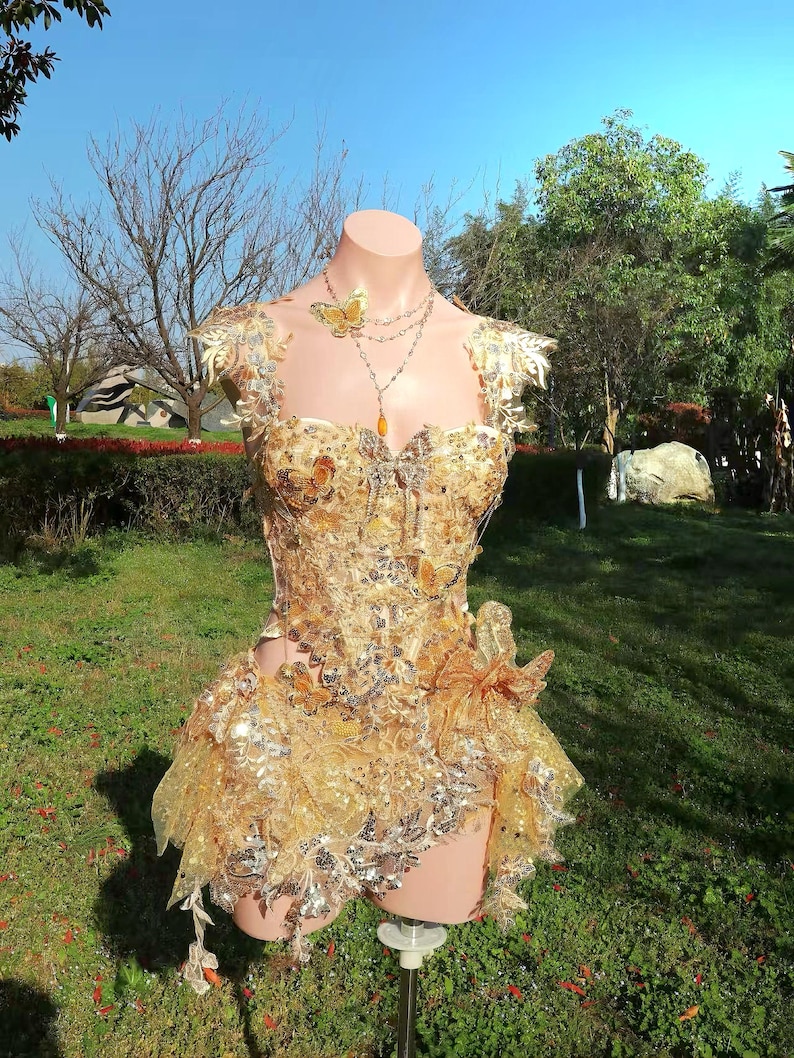 May include: A golden, embellished bodice and skirt ensemble, adorned with butterfly accents and shimmering details. The outfit is displayed on a mannequin, showcasing its intricate design and textures. A delicate necklace with a butterfly pendant completes the look.