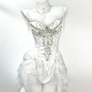 May include: A white corset-style dress with intricate silver beading and embellishments. The dress features a sweetheart neckline, delicate straps, and a layered tulle skirt. A matching choker necklace with a pendant and beaded shoulder accents complete the elegant design.