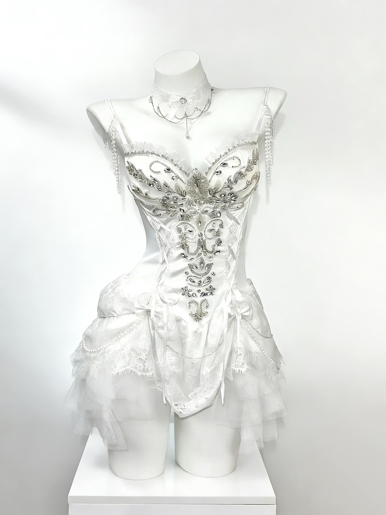 May include: A white corset-style dress with intricate silver beading and embellishments. The dress features a sweetheart neckline, delicate straps, and a layered tulle skirt. A matching choker necklace with a pendant and beaded shoulder accents complete the elegant design.