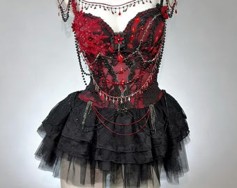 May include: A red and black gothic-style corset dress with a layered skirt, displayed on a mannequin. The bodice features intricate lace, bead, and jewel embellishments. A matching choker and beaded shoulder accents complete the ensemble.