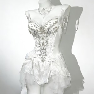 May include: A white corset-style dress with intricate silver embellishments and a layered skirt. The dress is displayed on a mannequin, featuring a matching choker and beaded accents. The bodice is detailed with floral and leaf patterns.