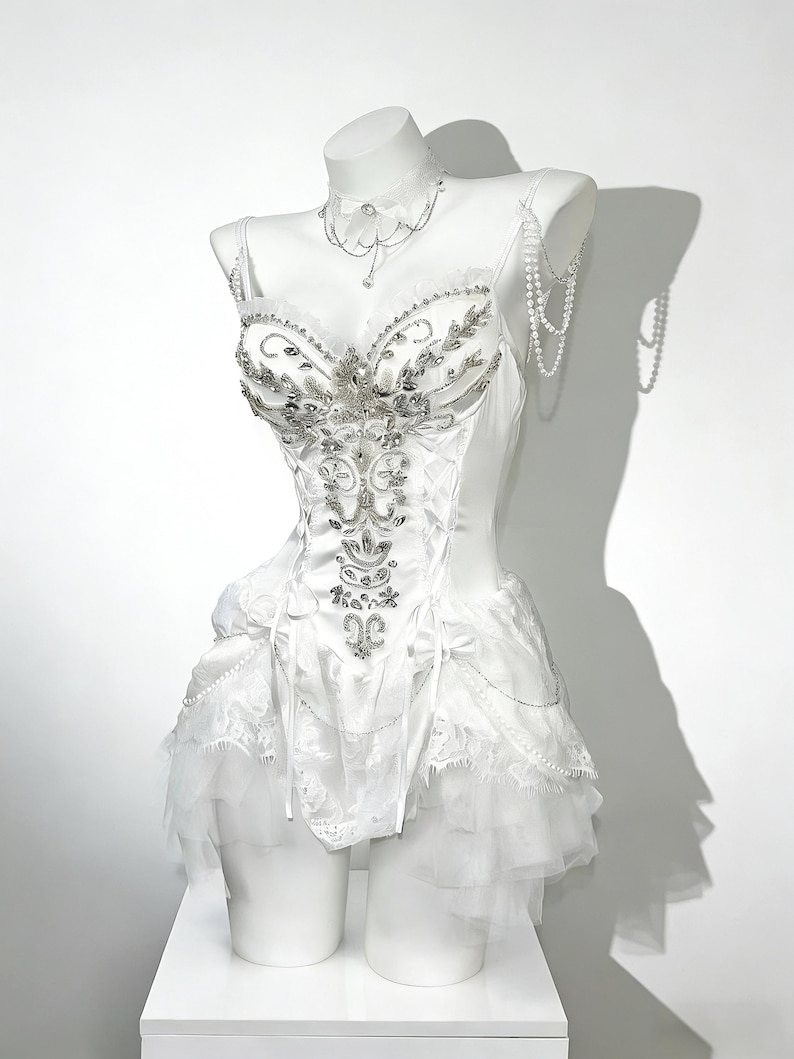 May include: A white corset-style dress with intricate silver embellishments and a layered skirt. The dress is displayed on a mannequin, featuring a matching choker and beaded accents. The bodice is detailed with floral and leaf patterns.