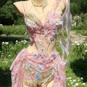 May include: A pink, embellished costume displayed on a mannequin. The outfit features a bodice with floral and beaded accents, a skirt, and a flowing ribbon. The costume is adorned with pearls, sequins, and blue accents, creating a whimsical fairy-like aesthetic.
