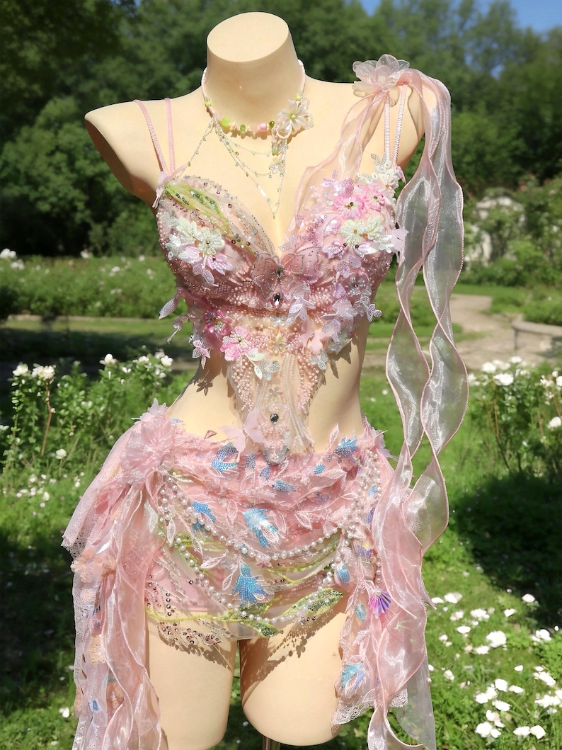 May include: A pink, embellished costume displayed on a mannequin. The outfit features a bodice with floral and beaded accents, a skirt, and a flowing ribbon. The costume is adorned with pearls, sequins, and blue accents, creating a whimsical fairy-like aesthetic.