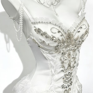 May include: A white satin corset-style top with intricate silver beading and embellishments. The top features a lace-up back, a ruffled neckline, and delicate pearl straps. A matching lace choker with a bow and dangling chains completes the elegant design.
