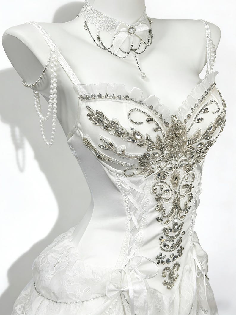 May include: A white satin corset-style top with intricate silver beading and embellishments. The top features a lace-up back, a ruffled neckline, and delicate pearl straps. A matching lace choker with a bow and dangling chains completes the elegant design.