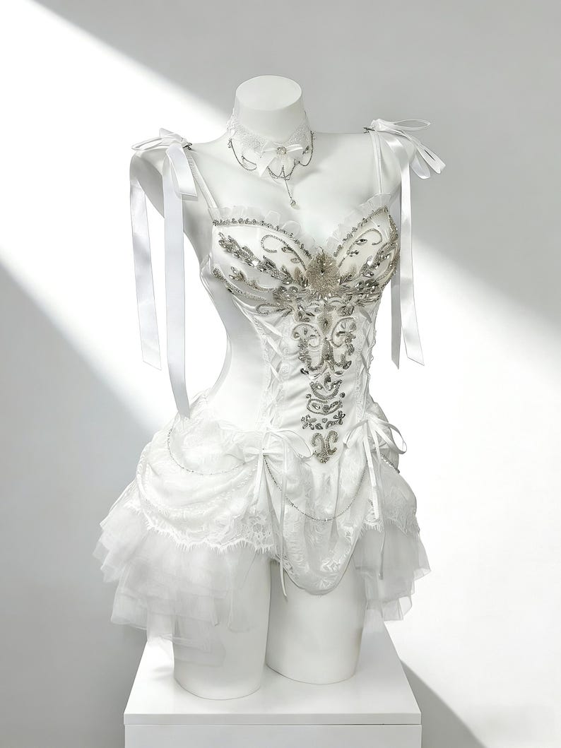 May include: White corset-style dress with silver embellishments and a layered skirt. The dress features ribbon straps and a matching choker necklace. The bodice is intricately detailed with silver beading and embroidery, creating a glamorous look.