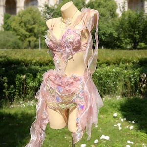 May include: A pink fairy costume displayed on a mannequin. The costume features a detailed bodice with floral embellishments, a layered skirt, and flowing ribbons. Iridescent accents and delicate beadwork add to the ethereal design. The costume is displayed outdoors.