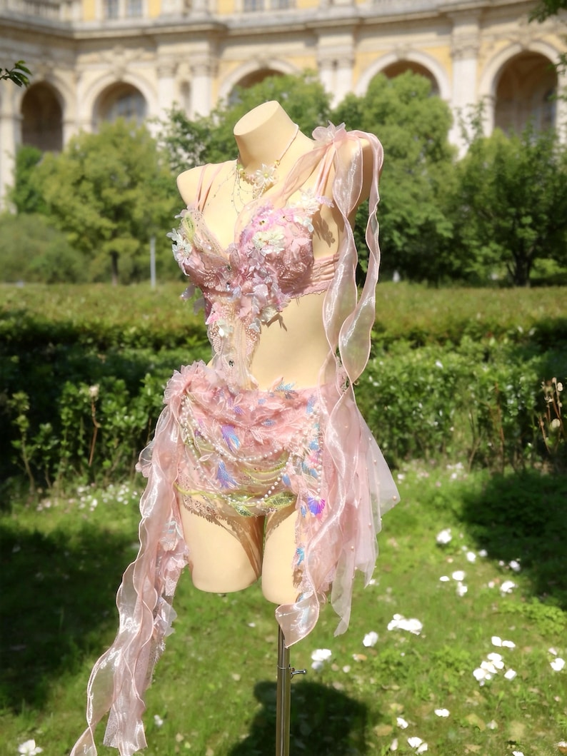 May include: A pink fairy costume displayed on a mannequin. The costume features a detailed bodice with floral embellishments, a layered skirt, and flowing ribbons. Iridescent accents and delicate beadwork add to the ethereal design. The costume is displayed outdoors.