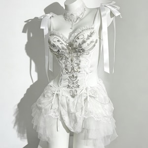 May include: A white corset-style dress with intricate silver embellishments and a layered skirt. The dress features delicate straps, bows, and a matching choker necklace. The dress is displayed on a white mannequin.