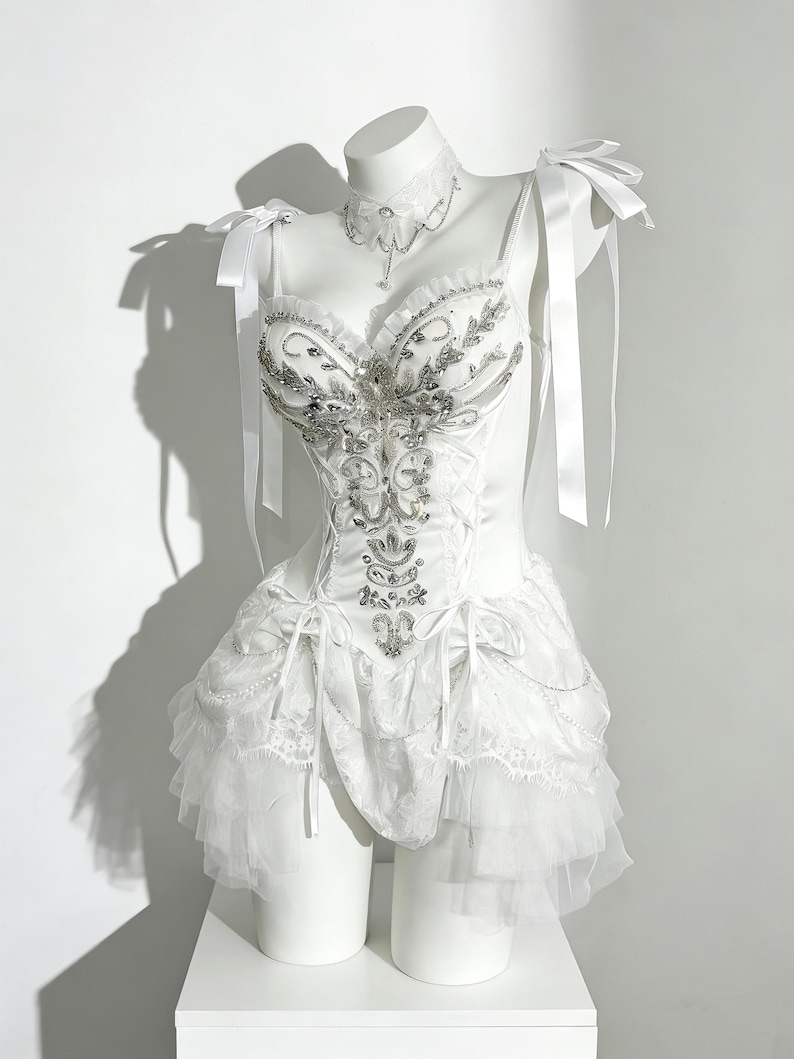 May include: A white corset-style dress with intricate silver embellishments and a layered skirt. The dress features delicate straps, bows, and a matching choker necklace. The dress is displayed on a white mannequin.