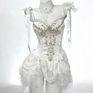 May include: A white corset-style dress with silver embellishments and a layered skirt. The dress features delicate straps, bows, and a matching choker necklace. The dress is displayed on a mannequin.