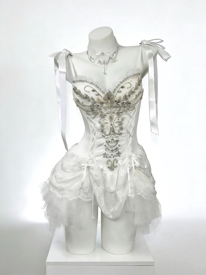 May include: A white corset-style dress with silver embellishments and a layered skirt. The dress features delicate straps, bows, and a matching choker necklace. The dress is displayed on a mannequin.