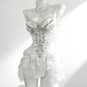 May include: A white corset-style dress with intricate silver beading and embellishments. The dress features a lace-up front, delicate straps, and a layered skirt. A matching choker and pearl strands adorn the mannequin, creating an elegant, formal look.