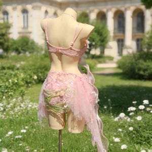 May include: A light pink lingerie set on a mannequin. The set includes a lace bra and a skirt with tulle and floral accents. The background features a building and a garden with white flowers.