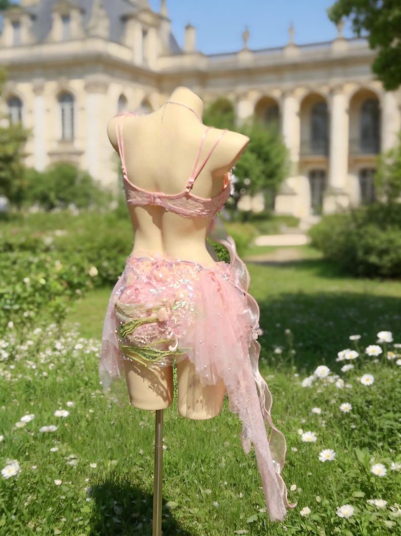 May include: A light pink lingerie set on a mannequin. The set includes a lace bra and a skirt with tulle and floral accents. The background features a building and a garden with white flowers.