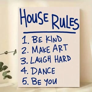 May include: A white rectangular sign with the words "HOUSE RULES" in blue script above a list of five rules: "BE KIND", "MAKE ART", "LAUGH HARD", "DANCE", and "BE YOU". The text is centered on the sign.