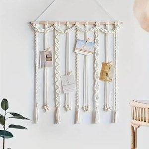 May include: A cream-colored macrame wall hanging with a wooden dowel and hanging string. The wall decor features multiple strands with various knotting patterns, holding small photos with wooden clips. The design includes tassels and decorative elements.